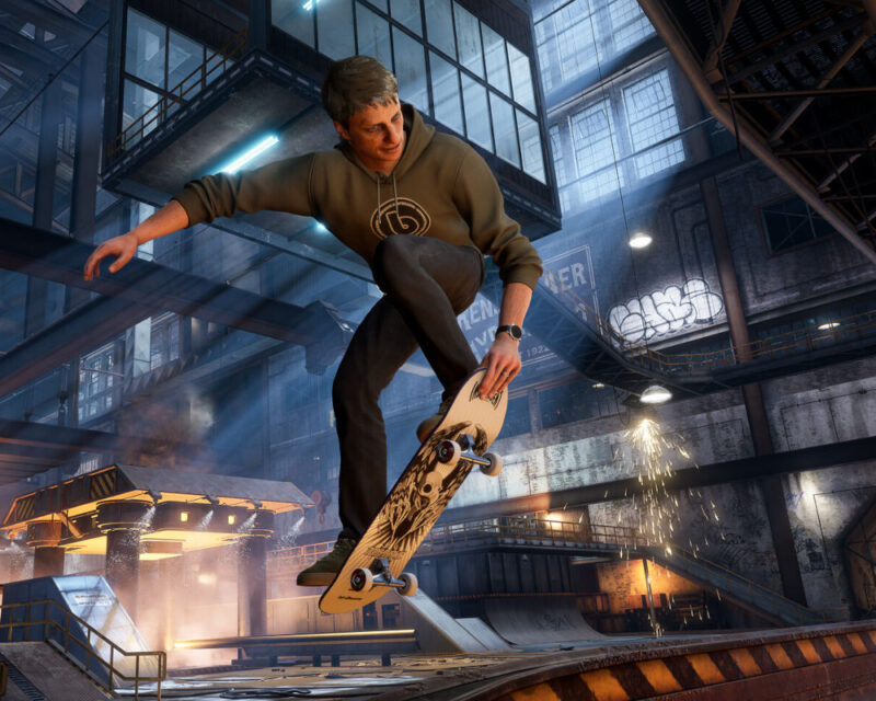 Na obrazku znajduje się Tony Hawk wykonujący trick na deskorolce. Jedną ręką trzyma przód deskorolki, a drugą ma uniesioną w górę. Znajduje się wewnątrz ciemnego budynku z dużą ilością okien.