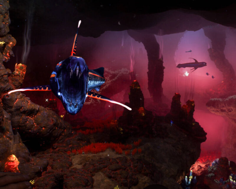 Na tym obrazku widzimy podwodną scenę, która wygląda jak kadr z gry wideo, z serii Subnautica. W centrum uwagi znajduje się ogromne, agresywnie wyglądające stworzenie morskie z otwartą paszczą pełną ostrych zębów. Jego ciało jest ciemnoniebieskie z jaskrawymi, pomarańczowymi i białymi bioluminescencyjnymi elementami. Stwór ten pływa w ciemnym, jaskiniowym środowisku. Wokół niego widać skaliste formacje o czerwonawo-pomarańczowej poświacie, sugerujące obecność lawy lub innego źródła ciepła i światła. W tle majaczą inne, mniejsze stworzenia morskie oraz dalsze fragmenty podwodnych jaskiń. Całość tworzy intensywną i potencjalnie niebezpieczną atmosferę eksploracji głębin oceanu.