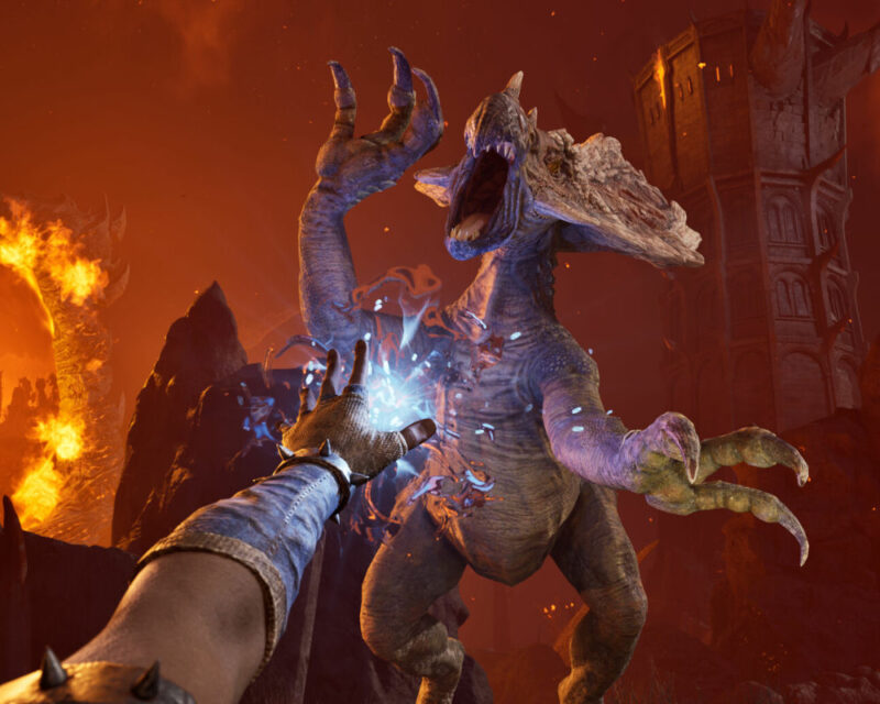 Na tym obrazku widzimy scenę z perspektywy pierwszej osoby, która wygląda jak fragment rozgrywki w grę fantasy. W centrum kadru znajduje się potężny, dwunożny stwór o szarej, łuskowatej skórze, z dużymi, ostrymi pazurami i otwartą paszczą, z której widać rzędy zębów. Jego głowa jest ozdobiona kostnymi wyrostkami. Przed nim, po lewej stronie, widzimy rękę w rękawicy z niebieskimi zdobieniami i złotymi detalami na nadgarstku. Z dłoni tej ręki emanuje jasnoniebieska, wirująca energia, sugerująca użycie magii lub jakiejś specjalnej mocy. Tło jest mroczne i ogniste. Po lewej stronie widać płomienie i ciemne, skaliste formacje. Po prawej stronie wznosi się fragment mrocznej, gotyckiej architektury, przypominającej zrujnowaną wieżę lub zamek, oświetloną od dołu przez pomarańczowe światło. Całość utrzymana jest w ciepłej, czerwono-pomarańczowej tonacji, kontrastującej z niebieską energią.