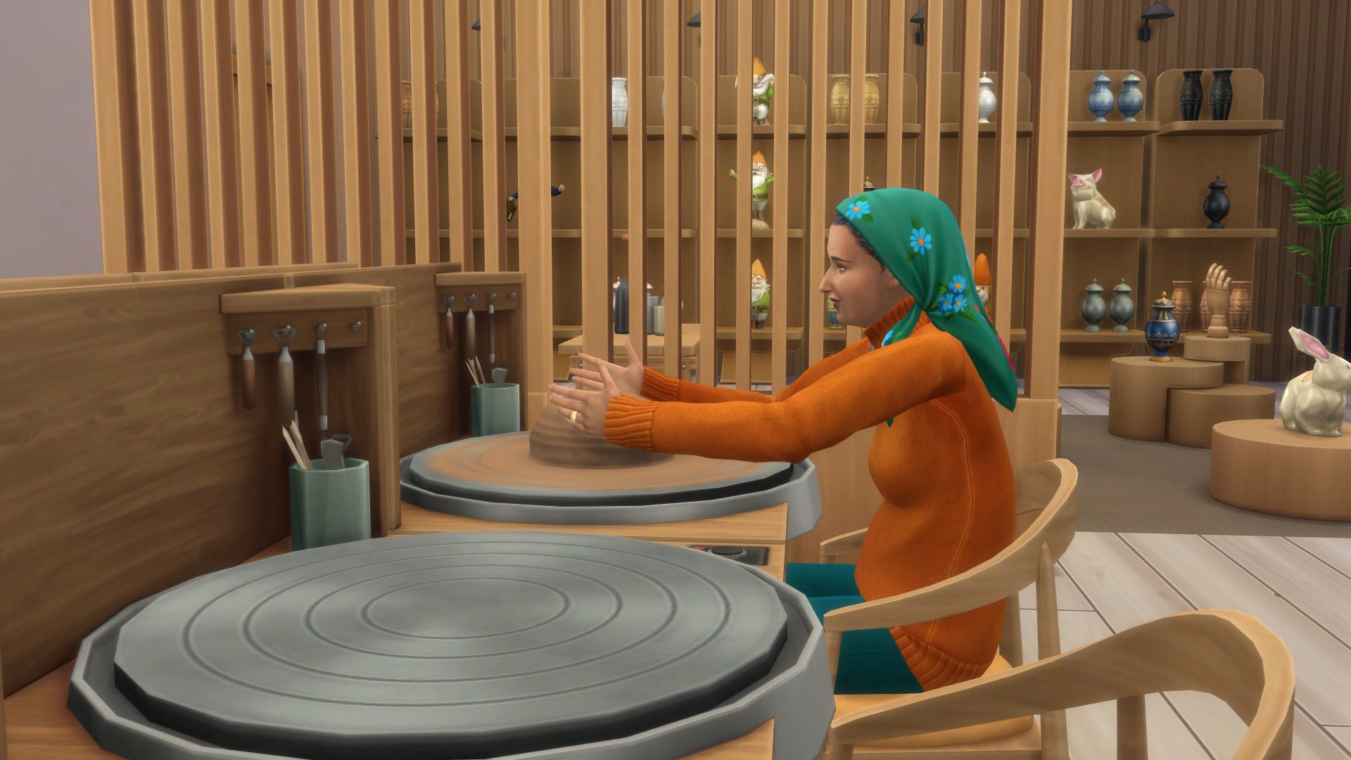 Zrzut ekranu z gry The Sims 4: Dochodowe hobby przedstawiający Simkę siedzącą przy kole garncarskim w pracowni ceramiki. Kobieta ma na sobie pomarańczowy sweter i zieloną chustę z niebieskimi kwiatami. Formuje gliniane naczynie na kole. W tle widać drewniane półki z gotowymi wyrobami ceramicznymi, takimi jak wazony, figurki i urny, a także dekoracyjne krasnale i roślinę doniczkową.