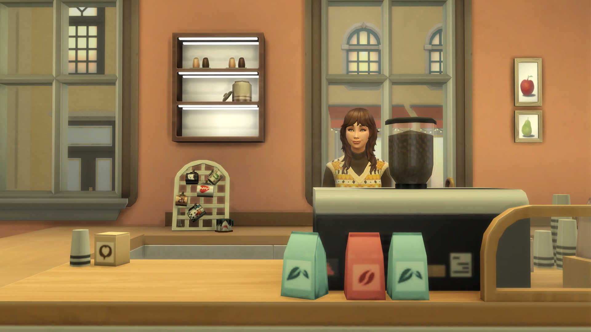 Zrzut ekranu z gry The Sims 4 przedstawiający baristkę stojącą za ladą w przytulnej kawiarni. Kobieta uśmiecha się, ubrana w żółty sweter w romby i brązowy golf. Przed nią znajduje się ekspres do kawy, trzy opakowania kawy oraz inne akcesoria baristyczne. W tle widać filiżanki na półkach, ozdobne obrazki owoców na ścianie oraz duże okna z widokiem na ulicę.