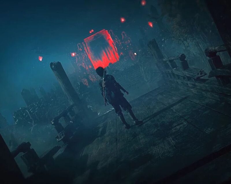 Na obrazku widzimy mroczną, klimatyczną scenę z gry — samotna postać w stroju przypominającym samuraja stoi na starym, zniszczonym moście, prowadzącym do ogromnych, świecących czerwienią wrót. Czerwona poświata oświetla mglistą, ponurą okolicę, pełną zarośli i ciemnych sylwetek budynków w tle. W powietrzu unoszą się czerwone latarnie lub świetliste punkty, potęgując wrażenie tajemniczości i zagrożenia. Cała scena przywodzi na myśl horror lub nadnaturalną przygodę osadzoną w feudalnej Japonii.