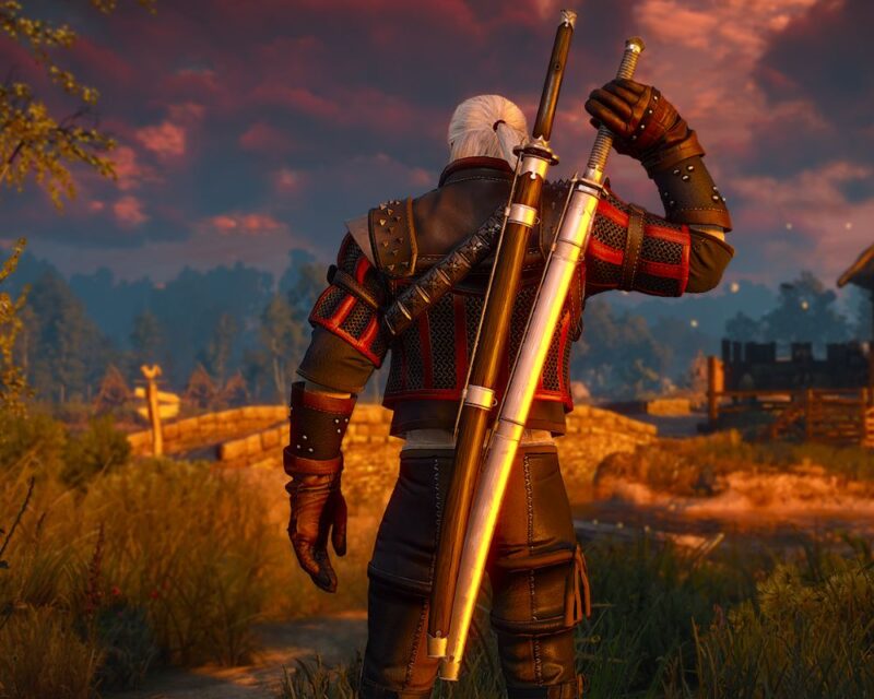 Postać wiedźmina Geralta z Rivii widziana od tyłu, stojąca na polnej drodze o zachodzie słońca. Ma białe włosy spięte w kucyk, skórzaną zbroję z czerwonymi akcentami oraz dwa miecze na plecach — stalowy i srebrny. W tle widać wieś, drewniany mostek, drzewa i purpurowe niebo pokryte chmurami. Scena pochodzi z gry „Wiedźmin 3: Dziki Gon” i oddaje epicki, fantastyczny klimat świata przedstawionego.