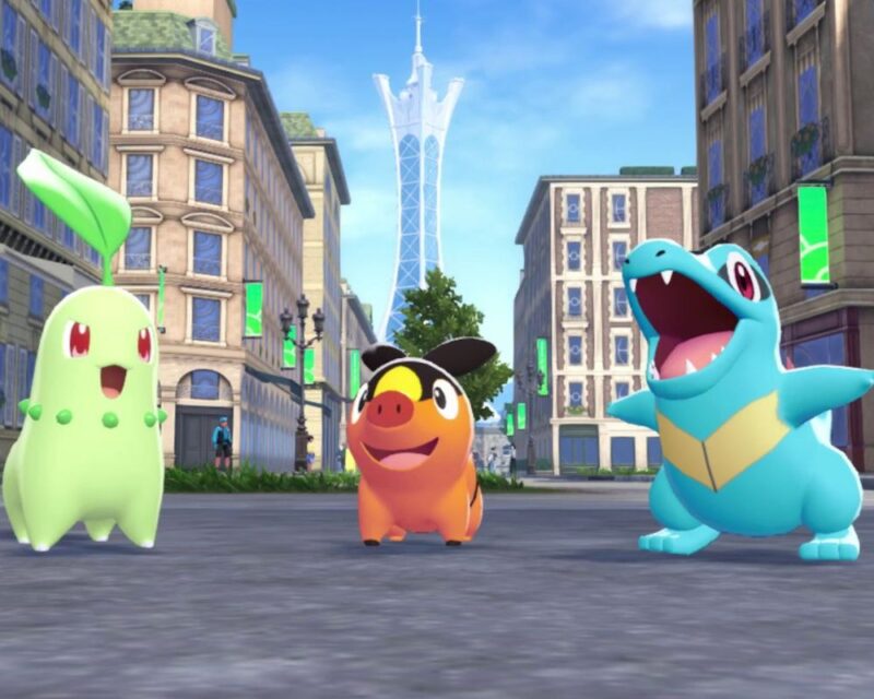 Na obrazku widzimy trzy kolorowe postacie z gry stojące na ulicy w mieście. Po lewej znajduje się zielony stworek z liściem na głowie, pośrodku pomarańczowy z czarnymi uszami, a po prawej niebieski z dużym uśmiechem i otwartą paszczą. W tle widoczne są wysokie budynki i wieża.