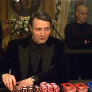 Na zdjęciu jest Mads Mikkelsen w roli Le Chiffre'a z Casino Royale siedzący przy pokerowym stole w kasynie.