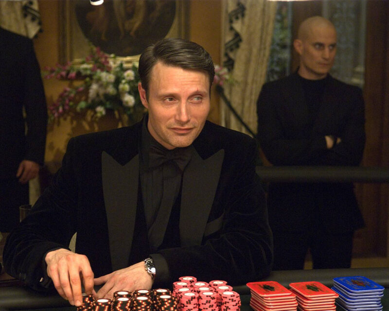 Na zdjęciu jest Mads Mikkelsen w roli Le Chiffre'a z Casino Royale siedzący przy pokerowym stole w kasynie.