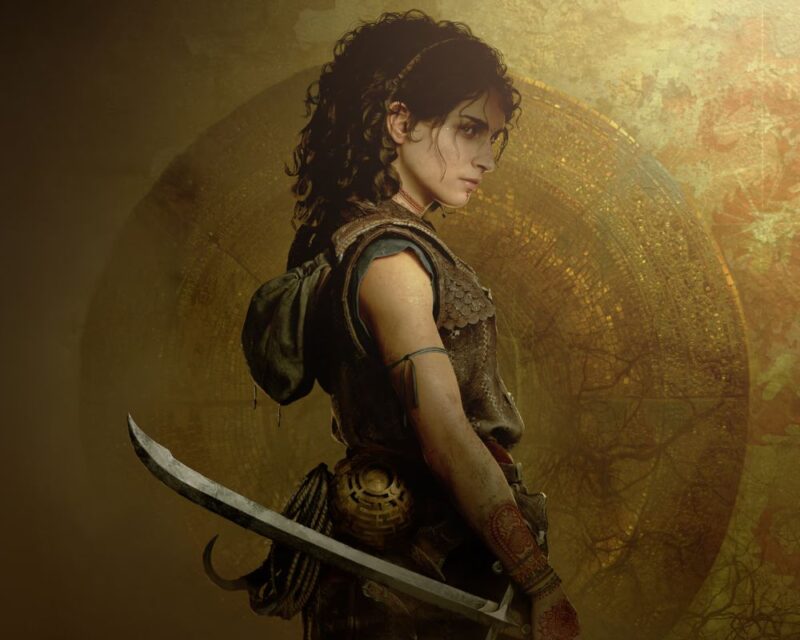 Na obrazku widać kobietę o ciemnych, kręconych włosach, ubraną w skórzaną zbroję. Stoi bokiem, trzymając zakrzywiony miecz, a na jej ramieniu widać opaskę.