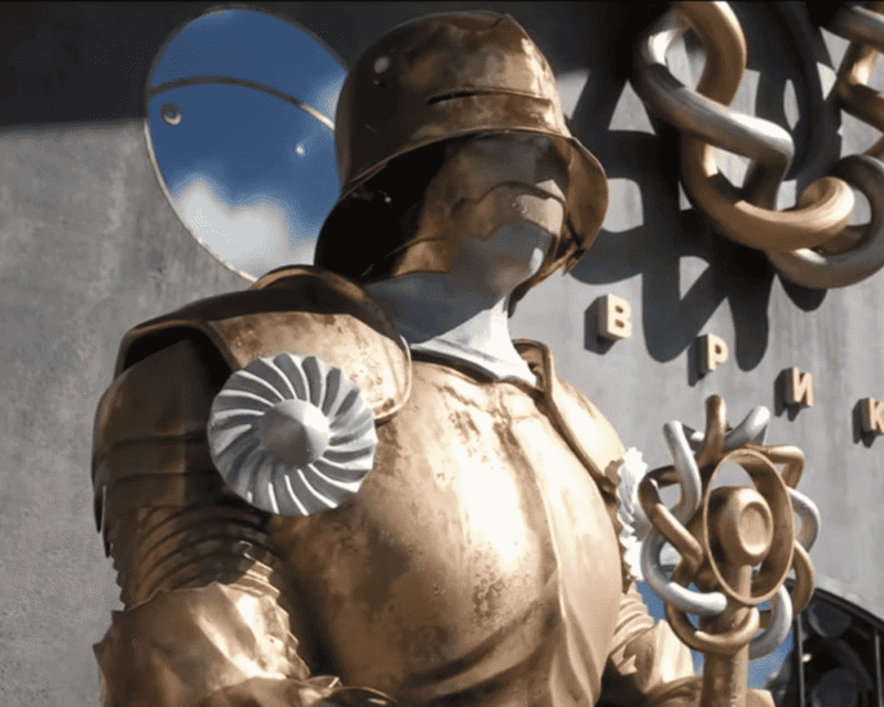 Zrzut ekranu z gry Atomic Heart 2 przedstawiający posąg retrofuturystycznego robota w złocistej zbroi, przypominającej styl radzieckiego konstruktywizmu. Postać trzyma symboliczną laskę z mechanicznym ornamentem, a na tle widać fragment budynku z cyrylicznym napisem. Styl wizualny typowy dla estetyki gry: połączenie brutalizmu, propagandy i surrealizmu technologicznego.