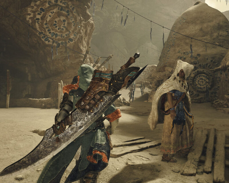 W centrum kadru, odwrócona plecami do widza, znajduje się postać gracza. Jest ona ubrana w solidną zbroję w odcieniach zieleni, brązu i błękitu, z elementami przypominającymi futro lub skórę. Na plecach postaci wisi imponujący, olbrzymi miecz, typowy dla gier Monster Hunter, z szerokim ostrzem i wyraźnymi detalami. Postać wydaje się być w ruchu, choć jej poza jest statyczna. Po prawej stronie obrazka, nieco dalej od postaci gracza, stoi inna postać, ubrana w jasne, postrzępione szaty z kapturem, które zakrywają większość jej twarzy. Szaty te są w odcieniach beżu i bieli, z niebieskimi i brązowymi detalami. Tło sceny to skalisty krajobraz, przypominający kanion lub jaskinię. Ściany skalne są beżowe i brązowe, z widocznymi teksturami i rzeźbieniami. Na jednej ze ścian w tle widać duży, okrągły wzór. Z sufitu skalnego zwisają liczne, podłużne obiekty, być może dekoracje lub formacje skalne, przypominające kryształy lub wiszące ozdoby. Całość sceny jest oświetlona w sposób sugerujący światło dzienne wpadające do wnętrza. Ogólny nastrój obrazka jest nieco tajemniczy i przygodowy, typowy dla gier fantasy i RPG.