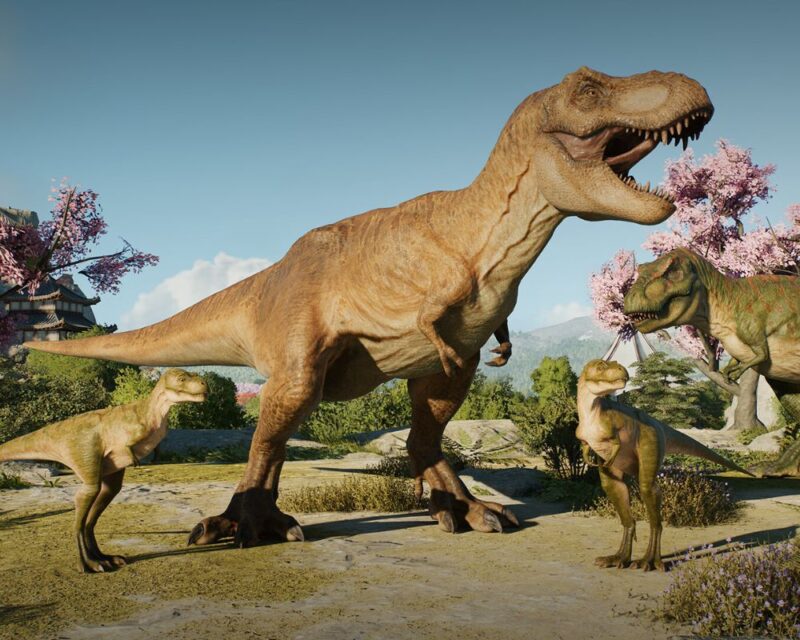 Na obrazku widzimy realistycznie odwzorowaną scenę z udziałem dinozaurów, z gry Jurassic World Evolution 3. Centralną postacią jest ogromny tyranozaur z otwartą paszczą, otoczony przez mniejsze dinozaury, które wyglądają na młode osobniki tego samego gatunku. W tle widać bujną, zieloną roślinność, góry i kwitnące drzewa wiśniowe, nadające scenerii niemal sielankowy charakter. Po lewej stronie w oddali można dostrzec fragment tradycyjnej japońskiej architektury, co nadaje całej kompozycji unikalnego klimatu. Scena łączy prehistoryczną faunę z subtelnym, orientalnym otoczeniem, sugerując nową, zaskakującą lokalizację w serii gier o dinozaurach.