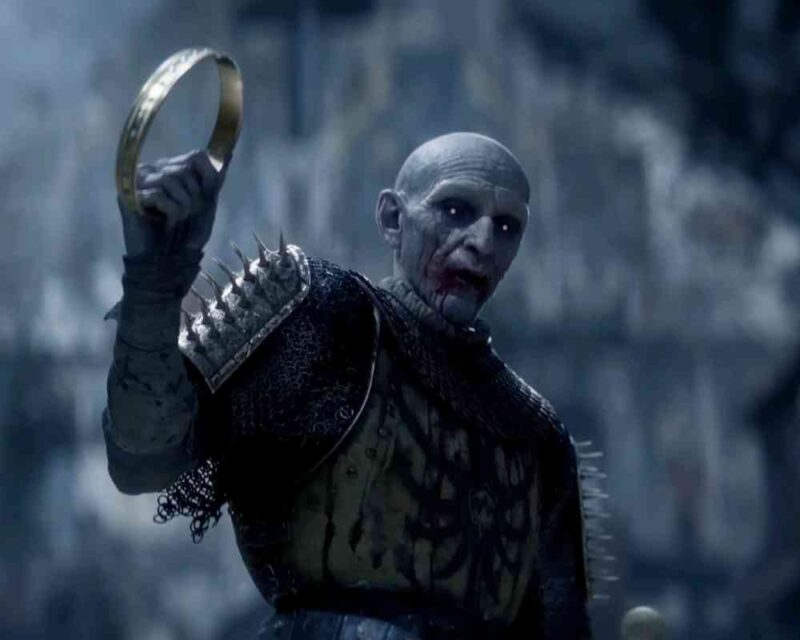 Postać z gry The Blood of Dawnwalker — blady, upiorny wojownik o łysiej głowie i czerwonych oczach, ubrany w zbroję z kolczastymi naramiennikami, trzymający w uniesionej ręce złoty pierścień. Tło jest ciemne i zamglone, podkreślając mroczny klimat sceny.