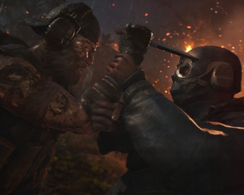 zrzut ekranu z gry Tom Clancy's Ghost Recon Breakpoint, widać na nim dwie walczące postaci, po lewej mężczyzna z gęstą brodą i bandaną, w prawej dłoni trzyma nóż i kieruje na napastnika, który przytrzymuje rękę swoimi obiema rękoma, jest ubrany w kombinezon, a twarz zakrywa maska