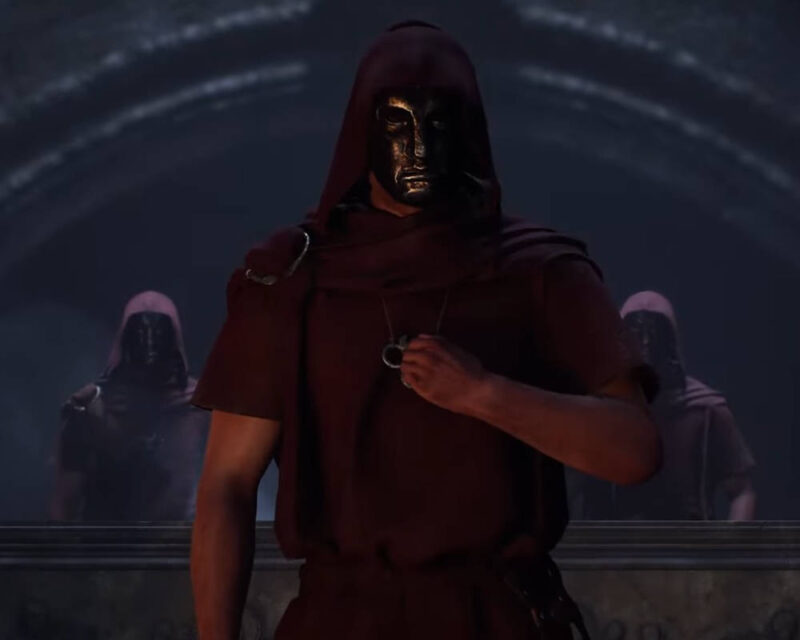 Na obrazku widać mężczyznę ubranego w bordową, luźną szatę z kapturem, która zakrywa jego głowę. Na twarzy ma on złocistą, błyszczącą maskę, która wydaje się mieć stylizowane, geometryczne rysy, z ledwo widocznymi otworami na oczy. Mężczyzna trzyma w prawej ręce mały, srebrny przedmiot przypominający pierścień lub medalion, który wisi na łańcuszku. Jego lewa ręka jest w opuszczona. Kostium jest pognieciony, co nadaje mu realistyczny wygląd. W tle, za główną postacią, widać dwie inne, nieco mniej ostre postacie, również ubrane w podobne, bordowe kaptury i maski. Stoją one w półcieniu, co nadaje scenie tajemniczy i rytualny charakter. Cała scena rozgrywa się w ciemnym, kamiennym pomieszczeniu, prawdopodobnie w jakiejś starożytnej świątyni lub grobowcu, z widocznymi w górnej części obrazu kamiennymi łukami lub kolumnami. Światło jest stonowane i dramatyczne, padające głównie na pierwszoplanową postać.