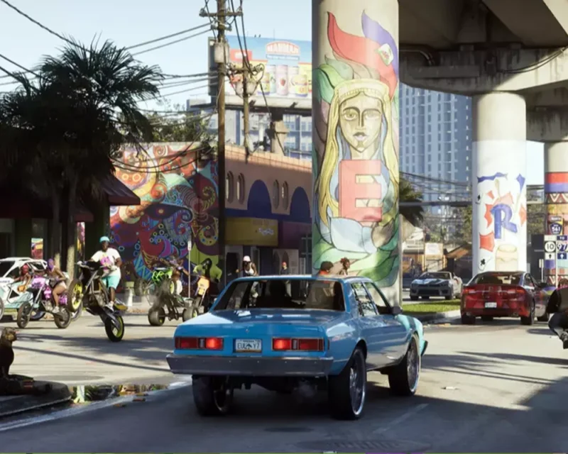 Ulica w Vice City w GTA VI – kolorowe murale na filarach, motocykliści i niebieski samochód w centrum kadru.