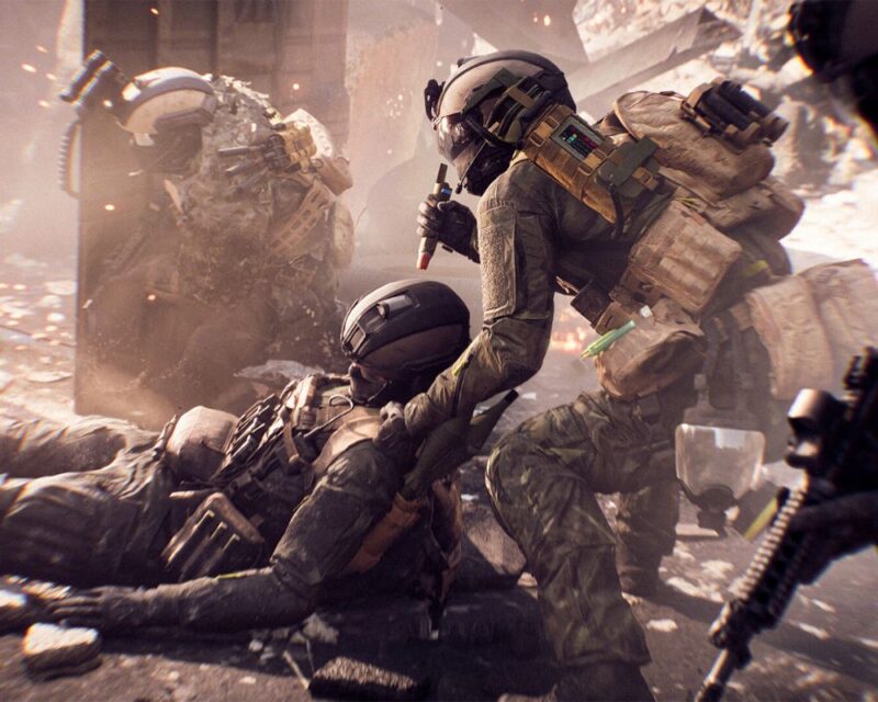 grafika z gry Battlefield 6, widzimy kilka postaci ubranych w kombinezony bojowe, na pierwszym planie znajdują się dwie, jedna leży, druga kuca nad nią i podtrzymuje jedną ręką, w drugiej trzyma strzykawkę, w tle kurz i ruiny budynków