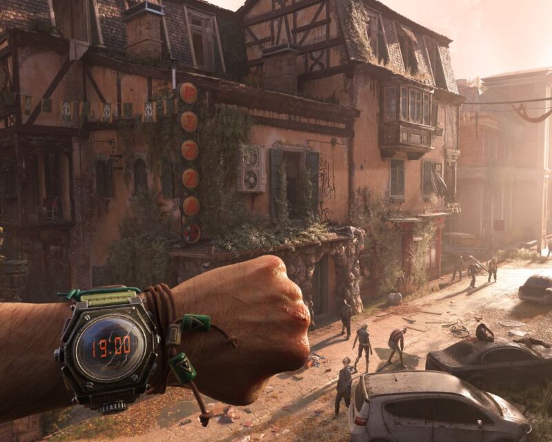 grafika z gry Dying Light: The Beast, przedstawia opuszczoną wioskę, widzimy podniszczone budynki i szyld z napisem PIZZA, litery idą od góry do dołu, niżej na ziemi widzimy chodzące zombie i wraki samochodów, na pierwszym tle natomiast widać męską lewą dłoń zaciśniętą w pięść i fragment ramienia, na nadgarstku natomiast jest zegarek z wyświetloną na czerwono liczbą 19:00