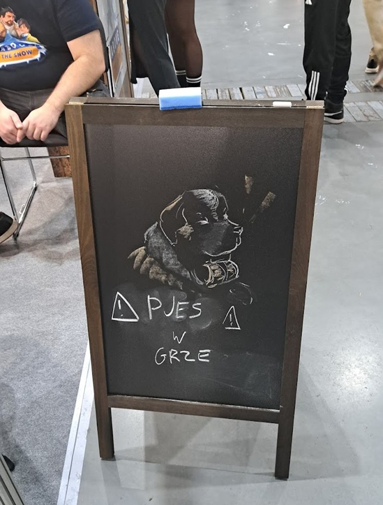 Drewniana tablica stojąca na podłodze hali targowej. Widnieje na niej rysunek psa w stylu chalk art oraz napis „Pies w grze” z ostrzegawczymi trójkątami. W tle osoby siedzące i przechodzące obok stoiska.
