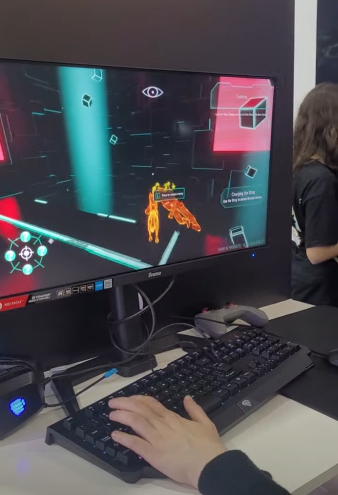 Osoba grająca na komputerze podczas targów gier. Na monitorze widać futurystyczny interfejs z neonowymi kolorami, postacią zaznaczoną na pomarańczowo i instrukcjami treningowymi. Na biurku klawiatura, myszka i kontroler.