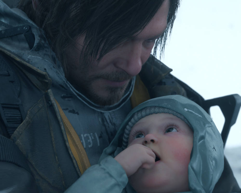 Na zbliżeniu widać Sama Portera Bridgesa (postać z gry Death Stranding, graną przez Normana Reedusa) trzymającego BB (Bridge Baby). Sam ma długie, ciemne włosy i patrzy na dziecko z łagodnym wyrazem twarzy. Jest ubrany w techniczny strój kuriera z żółtymi akcentami. Dziecko ma na sobie niebieską czapkę i kaptur, patrzy w górę, a jego rączka znajduje się przy ustach. Scena ma chłodną, szarobłękitną kolorystykę, sugerującą, że akcja dzieje się na zewnątrz.