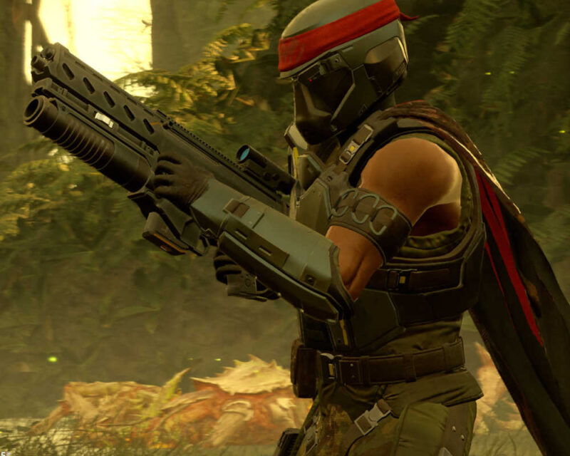 Na obrazku widać postać w zbroi i hełmie, która trzyma w rękach duży karabin lub strzelbę z podwójnymi lufami. Postać ma na sobie ciemnozielony lub czarny pancerz z czerwoną opaską/bandaną zawiązaną na czole pod hełmem oraz czerwoną pelerynę zarzuconą na ramiona. Ubiór i opaska nawiązują stylem do postaci Johna Rambo. Scena rozgrywa się w lesistym lub dżunglowym otoczeniu, z gęstą roślinnością i zamglonym, jasnym tłem, co sugeruje, że jest to kadr z gry wideo Helldivers 2. Postać jest skierowana w prawą stronę, w pozycji bojowej.