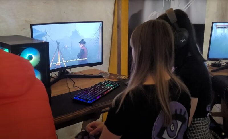 Dwie kobiety siedzące przy monitorze z grą. Na monitorze mężczyzna i kociołek nad ogniem.