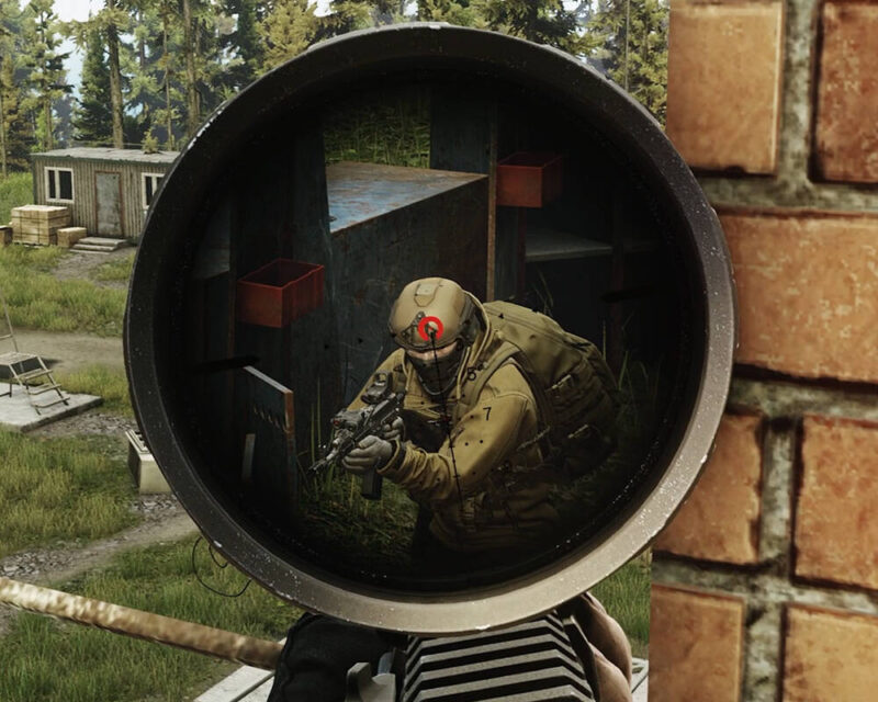 Obrazek przedstawia widok z celownika optycznego, prawdopodobnie karabinu snajperskiego, skupiony na uzbrojonym żołnierzu w terenie. Celownik ma charakterystyczną siatkę celowniczą z czerwonym punktem lub znakiem celowania umieszczonym na głowie żołnierza. Żołnierz jest ubrany w kamizelkę taktyczną i hełm, trzymając w rękach karabin szturmowy. Klęczy lub przykuca za osłoną wykonaną z ciemnych, geometrycznych bloków, w otoczeniu trawy i drzew. W tle widoczny jest teren leśny, jakiś mały drewniany budynek oraz czerwone, prostokątne pojemniki na półkach za celem. Na pierwszym planie, poza celownikiem, widoczna jest niewyraźna tekstura, która sugeruje dłoń i ramię osoby trzymającej broń. Cała scena ma militarny charakter i pochodzi z gry wideo, najprawdopodobniej jest to "Escape from Tarkov" ze względu na styl graficzny i detale wyposażenia.