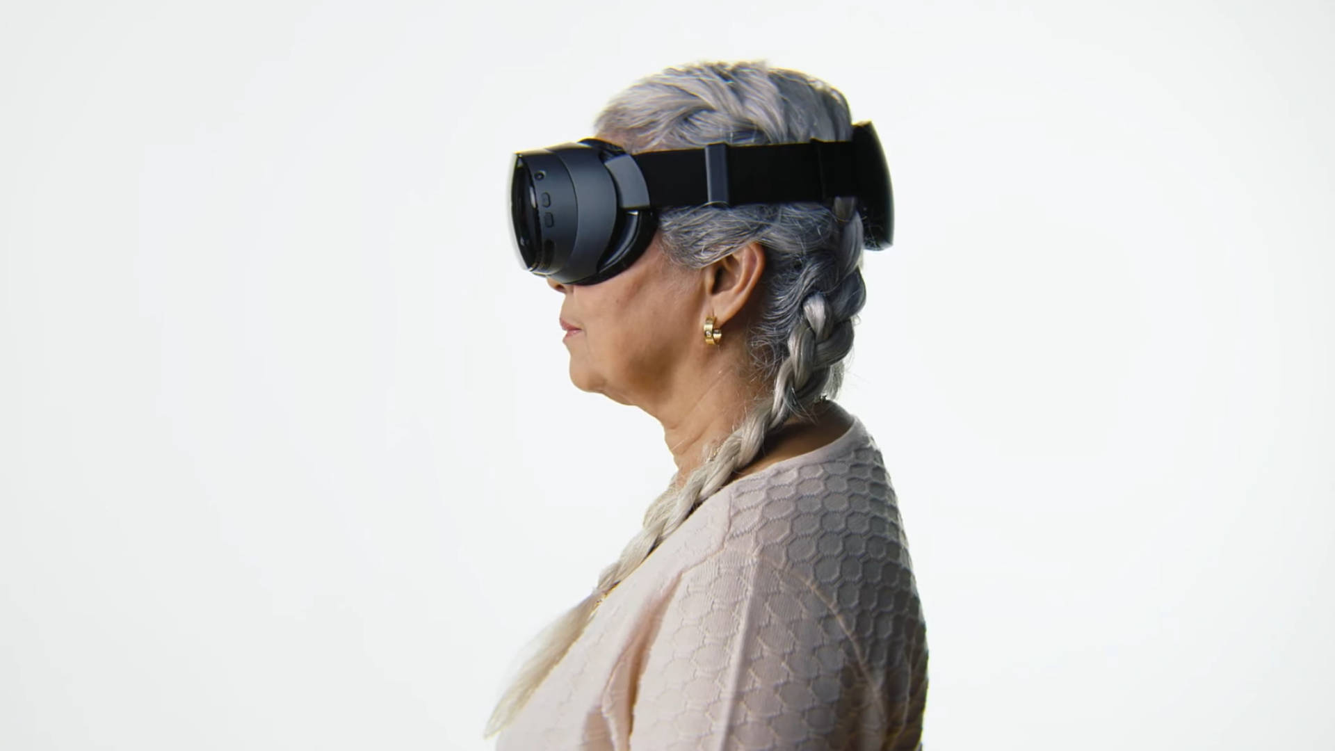 Na zdjęciu, uchwyconym z boku na wysokości głowy, widoczna jest starsza kobieta z siwymi włosami, splecionymi w warkocz, która ma na sobie czarne gogle wirtualnej rzeczywistości (VR), zakrywające jej oczy. Gogle, prawdopodobnie model Valve Index lub podobny, są przymocowane do głowy za pomocą czarnych pasków. Kobieta jest ubrana w jasny, beżowy lub bladoróżowy sweter. Ma na sobie mały, złoty kolczyk w uchu. Tło jest jednolicie białe i jasne, co mocno kontrastuje z ciemnymi goglami i skupia uwagę na twarzy i urządzeniu VR. Kobieta ma lekko zaciśnięte usta, sugerując skupienie na wirtualnym doświadczeniu.