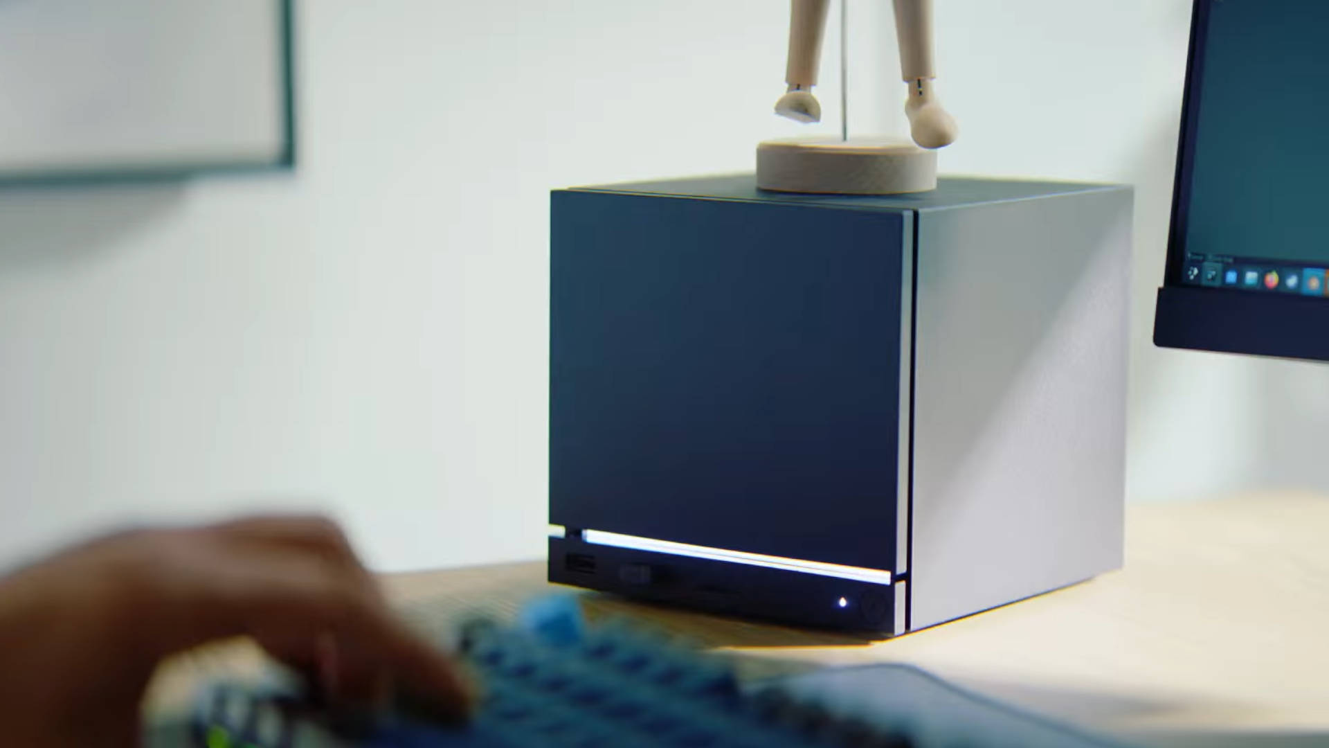 
Na zdjęciu widać mały komputer typu Steam Machine o kształcie prostopadłościanu w kolorach ciemnoszarym i srebrnym. Urządzenie stoi na jasnym, drewnianym biurku. Z przodu, u dołu ciemnej obudowy, znajduje się poziomy pasek z portami (prawdopodobnie USB) oraz mała dioda świecąca. Na górnej części obudowy umieszczony jest drewniany model manekina (prawdopodobnie artystyczny) z podniesionymi nogami, stojący na małej okrągłej podstawie. Po prawej stronie widoczny jest fragment czarnego monitora, na którego ekranie widać ikonę kosza na śmieci. W lewym dolnym rogu znajduje się fragment dłoni użytkownika oraz nieostra, kolorowa klawiatura mechaniczna, co sugeruje, że urządzenie jest w użyciu. Tło stanowi jasna ściana.