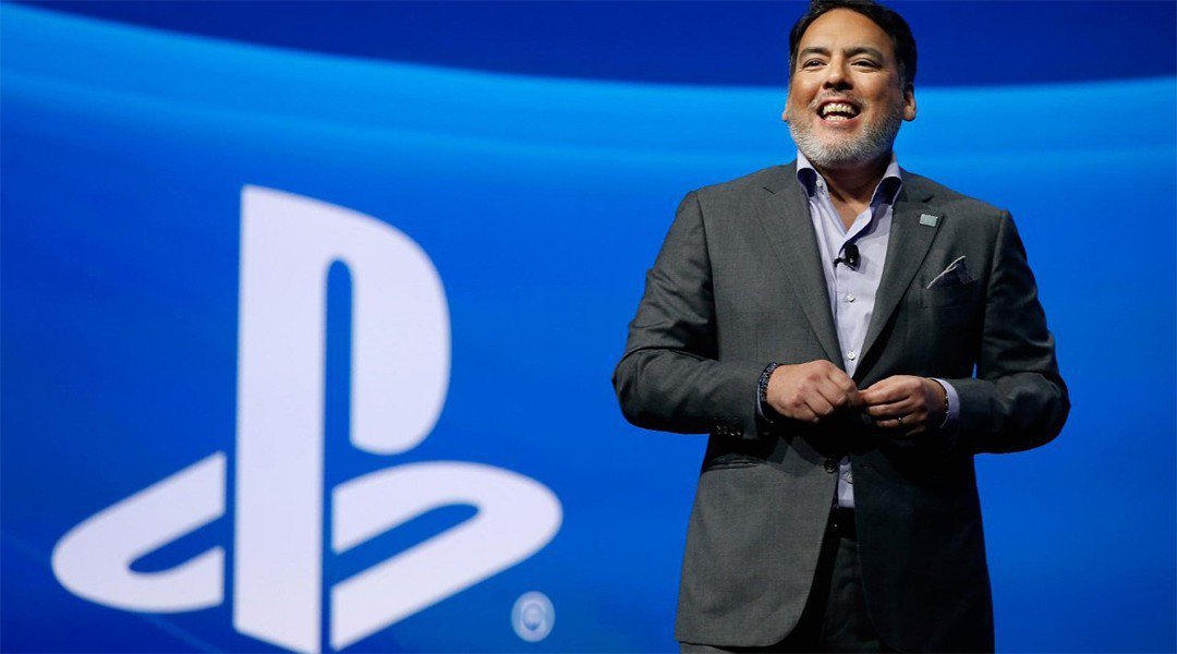 Na zdjęciu widzimy Shawna Laydena, Byłego szef Sony Interactive Entertainment / PlayStation Studios. Ubrany jest w garnitur i jest w trakcie jakiegoś przemówienia. Stoi na tle dużego niebieskiego banneru z logiem Playstation