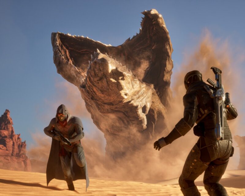 Grafika z gry Dune: Awakening przedstawia dwóch uzbrojonych bohaterów na pustyni Arrakis, którzy stają w obliczu gigantycznego czerwia pustyni wynurzającego się z piasku. Scena jest dynamiczna, pełna pyłu i napięcia, podkreślając niebezpieczeństwo świata inspirowanego uniwersum "Diuny".