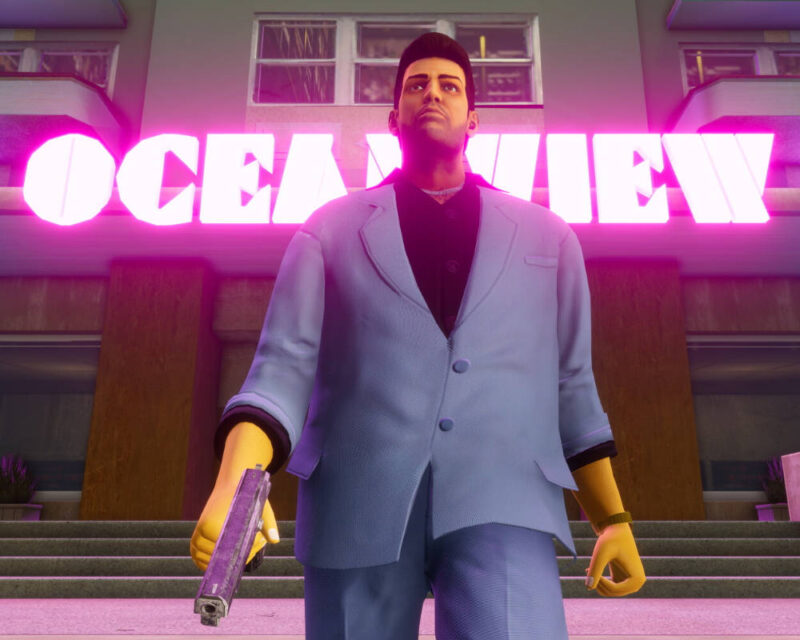 Na obrazku widać męską postać, Tommy’ego Vercettiego, głównego bohatera gry Grand Theft Auto: Vice City, stojącego na tle neonowego napisu „OCEAN VIEW” zdobiącego hotel o tej samej nazwie. Mężczyzna ma na sobie charakterystyczny błękitny garnitur z ciemną koszulą pod spodem, a w prawej dłoni trzyma pistolet skierowany lufą w stronę ziemi. Cała scena utrzymana jest w stylistyce retro lat 80., zdominowanej przez intensywne różowe i fioletowe światło neonów, co nadaje grafice kinowy i nostalgiczny klimat charakterystyczny dla estetyki synthwave. Postać patrzy lekko w górę z poważnym wyrazem twarzy, a tło stanowią schody prowadzące do hotelu oraz fragmenty fasady budynku.