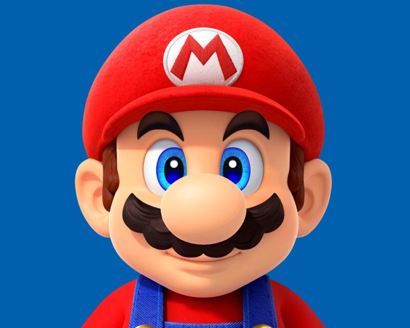 Zbliżenie na twarz Mario z Super Mario bros. WIdzimy najbardziej klasyczne wcielenie bohatera, w swoich niebieskich ogrodniczkach, czerwonej kolszulce i dużej czerwonej czapce z literą M na jej przodzie. Mario ma niebieskie oczy, duży nos i wąs. Stoi na niebieskim tle.