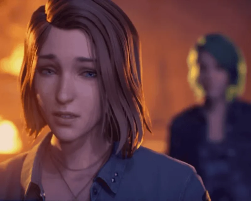 Zbliżenie na zapłakaną młodą kobietę o krótkich włosach, stojącą na tle ognia; w tle rozmyta sylwetka drugiej osoby. Scena z gry Life is Strange: Reunion