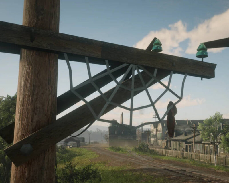 Na obrazku widać kadr z gry wideo, prawdopodobnie Red Dead Redemption 2, przedstawiający zbliżenie na drewniany słup telegraficzny, na którym rozpięta jest duża, nienaturalnie biała i geometryczna pajęczyna. Konstrukcja pajęczyny jest bardzo wyraźna i przypomina celowe oznaczenie, a w jej dolnej części zaplątane jest czerwonawe pióro lub kępka futra, co nadaje całości zagadkowy charakter. W tle rozciąga się widok na osadę o architekturze z przełomu XIX i XX wieku z drewnianymi budynkami i piaszczystą drogą biegnącą wzdłuż torów kolejowych. Niebo jest jasne z delikatnymi chmurami, a roślinność wokół miasteczka jest bujna i zielona, co sugeruje słoneczny dzień w świecie gry.