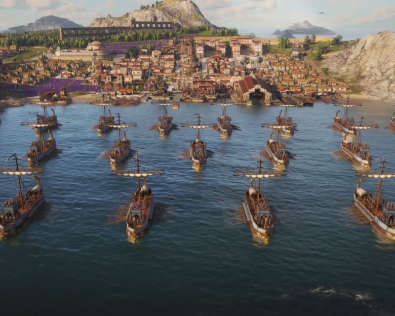 Grafika prezentuje scenę z gry Anno 117: Pax Romana. Widok od strony morza na flotę trirem oraz nabrzeże portu i rzymskie miasto. W tle widać akwedukt, góry i inne wyspy.