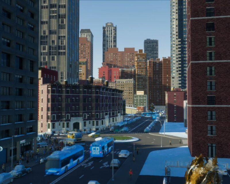 Obrazek prezentuje zrzut ekranu z gry Cities: Skylines 2 – zimowy krajobraz miejski, ruchliwa ulica z rondem i autobusami. Wzdłuż alei ciągną się kamienice i wieżowce. Na chodnikach sporo ludzi.
