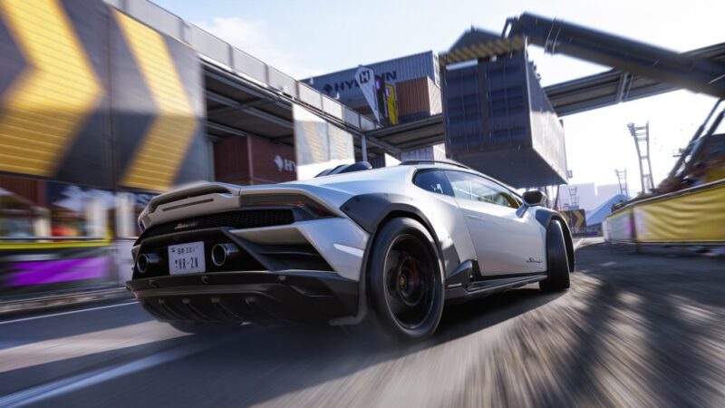 Co wiemy o Forza Horizon 6. Kadr przedstawia tył jasnego samochodu jadącego z dużą prędkością asfaltową drogą w obszarze przemysłowym, prawdopodobnie w dokach. Nad samochodem widać dźwig podnoszący kontener dużych rozmiarów.