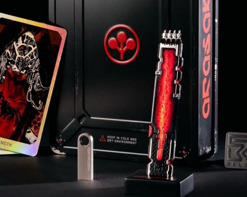 Zestaw kolekcjonerski Cyberpunk 2077 edycja kolekcjonerska Relic. Na zdjęciu widać metalowe, czarne opakowanie z logo Arasaki, podświetlaną replikę biochipa Relic na stojaku, trzy holograficzne karty tarota, pendrive z wygrawerowanym logo oraz dwie przypinki. Wszystkie elementy ułożone są na ciemnym tle, eksponując detale i futurystyczny design gadżetów.