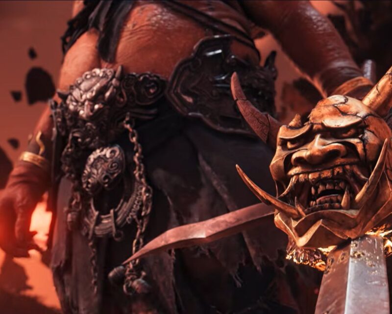 Zrzut ekranu ze zwiastuna gry Ghost of Yotei: Legends. Obraz prezentuje fragment wizerunku jednego z bossów – Oni – będącego ogropodobnym demonem z masywnymi kłami i rogami. W tym przypadku mamy rzut na dolną część postaci, odzianej w podartą, materiałową przepaskę z metalowym pasem, opatrzonym bogatą ornamentyką i łańcuchami. W dłoni trzyma miecz z jelcem prezentującym wyobrażenie Oni. W tle mroczny krajobraz z rdzawoczerwonym niebem i unoszącymi się fragmentami (najpewniej skałami).