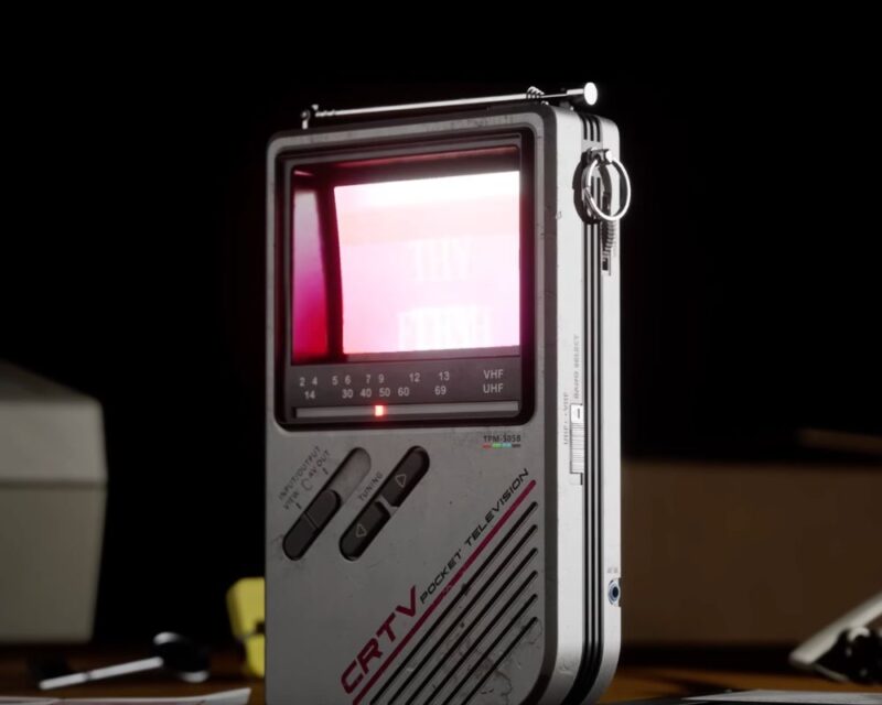 Zdjęcie przedstawia odbiornik krótkofalówki. Jest to szare, prostopadłościenne pudło z czerwonym wyświetlaczem, paskiem z widocznymi częstotliwościami, kilkoma przyciskami do regulacji dźwięku i anteną. Odbiornik stoi na biurku. W tle widzimy telefon, klucze i kilka niewyraźnych obiektów. Światło pada głównie na odbiornik, a wszystko za nim jest ciemne.