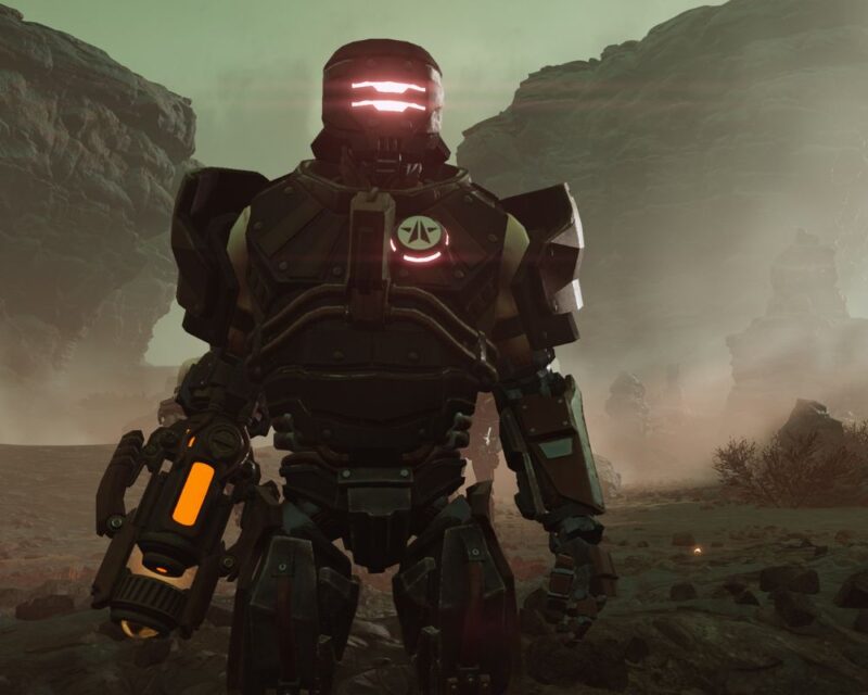 Grafika przedstawiająca cyborga w futurystycznej zbroi, ze świecącym hełmem, podświetlanym na czerwono oraz bronią z żółtymi elementami, zamiast ręki. Na zbroi znajduje się również białą odznaka z niebieską gwiazdą. W tle widać po oby stronach dwie skały oraz piach i krzaki.