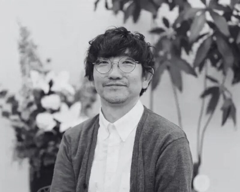 Na czarno-białym zdjęciu znajduje się mężczyzna w białej koszuli i szarym swetrze. Ma na sobie okulary, a jego włosy są ciemne, kręcone. W tle widać z lewej strony białe kwiaty, a po prawej stronie duża roślina z liśćmi, bez kwiatów