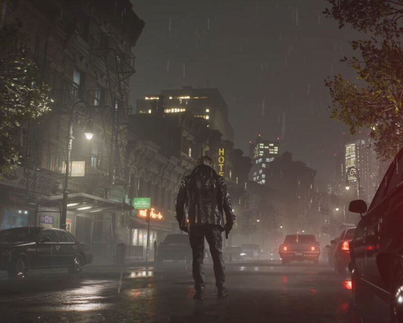 Zrzut ekranu z gry Resident Evil Requiem. Prezentuje nocną scenerię miasta w czasie deszczu. Leon S. Kennedy stoi na środku drogi tyłem do kamery, ubrany w ciemną skórzaną kurtkę i ciężkie buty. W prawej dłoni ma pistolet. Wkoło stoją samochody, latarnia uliczna rzuca miękkie światło. W tle widać szereg amerykańskich kamienic, a także nowoczesne wieżowce. Zrzut ekranu jednocześnie prezentuje skok technologiczny w zakresie oprawy wizualnej, osiągalnej na konsoli PS5 Pro.