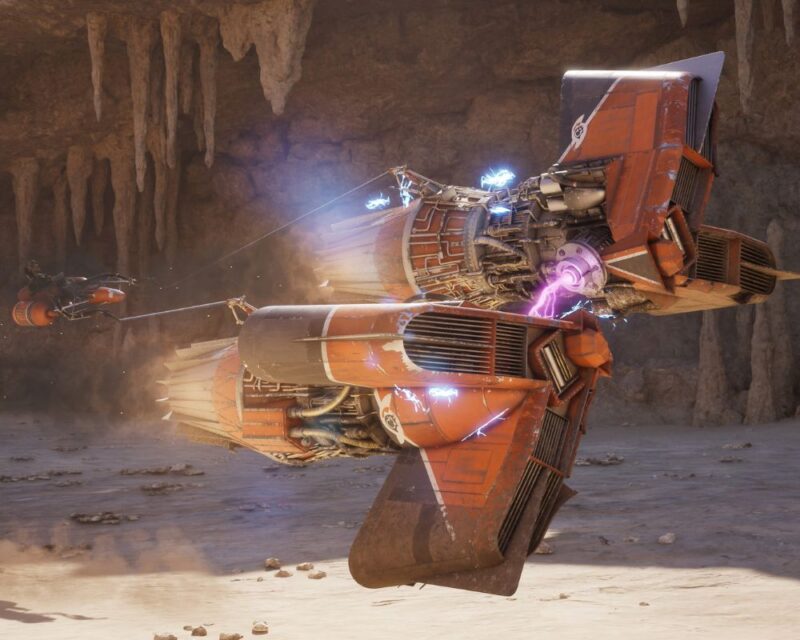 Czerwony, futurystyczny ścigacz typu podracer unosi się nad piaszczystym podłożem w skalnym kanionie. Z jego silników wydobywają się niebieskie iskry i fioletowe wyładowania elektryczne, a w tle widać drugi pojazd lecący na linie holowniczej. Wokół znajdują się wysokie, surowe ściany jaskini z wiszącymi formacjami skalnymi.