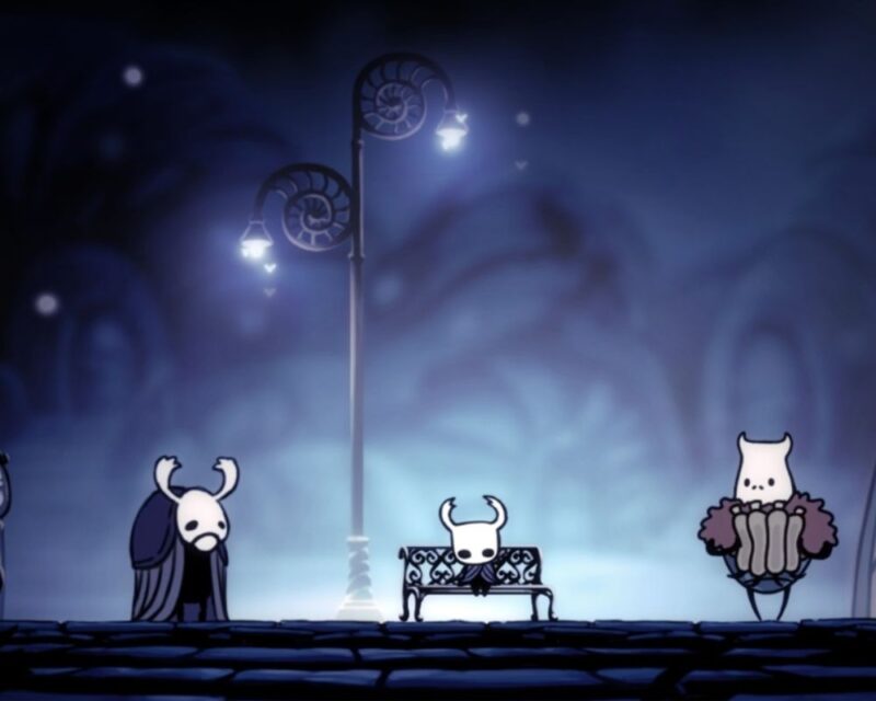 Na obrazie widać nastrojową scenę z gry Hollow Knight utrzymaną w chłodnej, niebieskiej kolorystyce. W centrum siedzi mała, rogata postać bohatera na ozdobnej ławce, pod delikatnie świecącą, stylizowaną latarnią uliczną. Po lewej stronie stoi zamyślona postać w długim płaszczu z opuszczoną głową, a po prawej znajduje się pulchny, biały bohater trzymający przed sobą mapę. Tło jest lekko rozmyte i przypomina spokojną, nocną uliczkę w fantastycznym, podziemnym mieście. Całość tworzy melancholijny, cichy klimat charakterystyczny dla świata gry.