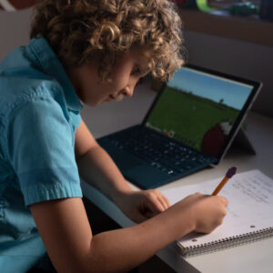 Młody chłopiec z kręconymi włosami siedzi przy biurku, pisząc w zeszycie i jednocześnie korzystając z laptopa z uruchomioną grą Minecraft. Zdjęcie przedstawia wykorzystanie nowoczesnych technologii w edukacji i nauce zdalnej.
