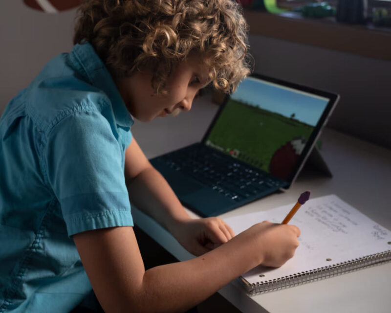 Młody chłopiec z kręconymi włosami siedzi przy biurku, pisząc w zeszycie i jednocześnie korzystając z laptopa z uruchomioną grą Minecraft. Zdjęcie przedstawia wykorzystanie nowoczesnych technologii w edukacji i nauce zdalnej.