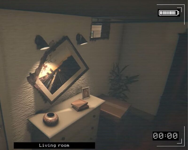 Obraz przedstawia niewielki fragment salonu widziany z perspektywy kamery monitoringu. Ujęcie ma lekko przyciemniony, ziarnisty wygląd, jak nagranie z kamery wideo z włączonym wskaźnikiem baterii w prawym górnym rogu i znacznikiem czasu „00:00” w prawym dolnym rogu. Na dole widnieje podpis „Living room”. Po lewej stronie stoi jasna komoda z trzema szufladami. Na jej blacie znajdują się trzy przedmioty: okrągły, dekoracyjny wazon w brązowo-miedzianym kolorze, stos dwóch książek oraz mała ramka ze zdjęciem. Nad komodą wisi duże, kwadratowe lustro zawieszone pod kątem – jest przekrzywione, jakby nie było równo zamocowane. Nad lustrem zamontowane są dwie czarne lampy ścienne skierowane w dół, oświetlające jego powierzchnię ciepłym światłem. W odbiciu lustra widać płomienie – wygląda to tak, jakby gdzieś w pomieszczeniu palił się ogień, choć sam ogień nie jest bezpośrednio widoczny poza odbiciem. Po prawej stronie komody stoi wysoka roślina doniczkowa o długich, wąskich liściach. W tle widać jasną ścianę oraz małe, prostokątne okno umieszczone wysoko pod sufitem. Cała scena ma spokojny, ale lekko niepokojący nastrój, głównie przez przekrzywione lustro i odbicie ognia.