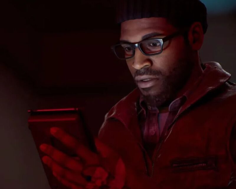 Simon Ordell, mężczyzna w okularach w średnim wieku ubrany w czerwoną kamizelkę i czarną czapkę trzymający telefon w dłoni, znajdujący się w ciemnym pomieszczeniu