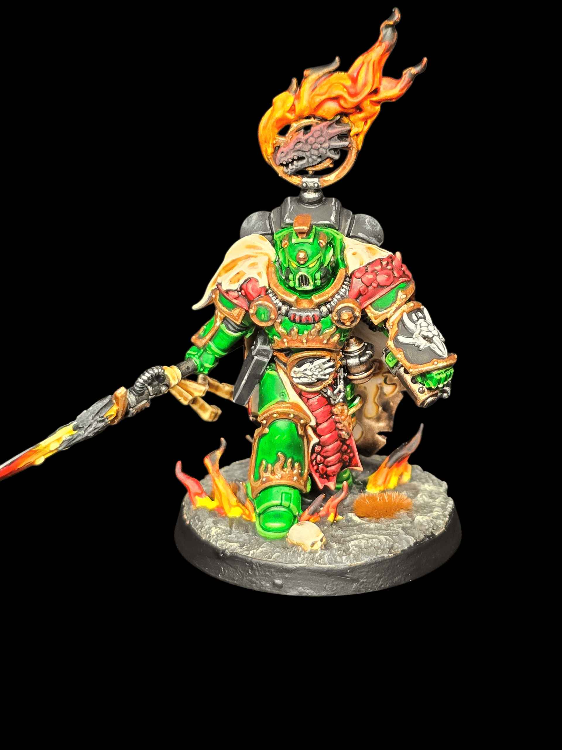 figurka z uniwersum Warhammer 40,000 Zdjęcie pomalowanej, kolekcjonerskiej figurki przedstawiającej futurystycznego wojownika (Space Marine) na czarnym tle. Postać zakuta jest w bogato zdobiony pancerz wspomagany o jaskrawozielonym kolorze ze złotymi krawędziami. Hełm ma świecące na czerwono wizjery, a naramienniki zdobią motywy smoczej/gadziej łuski. Nad plecakiem wojownika unosi się żelazna obręcz z głową smoka, z której buchają realistycznie pomalowane płomienie w kolorach żółci i pomarańczu. Postać trzyma włócznię z płonącym ostrzem. Figurka stoi na podstawce imitującej skalisty, popielaty teren, na którym również widać niewielkie płomienie i czaszkę.